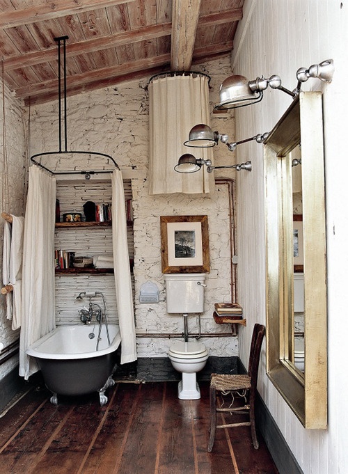 łazienka rustykalna w w typowo "wiejskim" stylu z kamieniami z ścianach, belkach na suficie i deskach na podłodze. Wolno stojąca żeliwna wanna i piękne duże w złotej ramie lustro